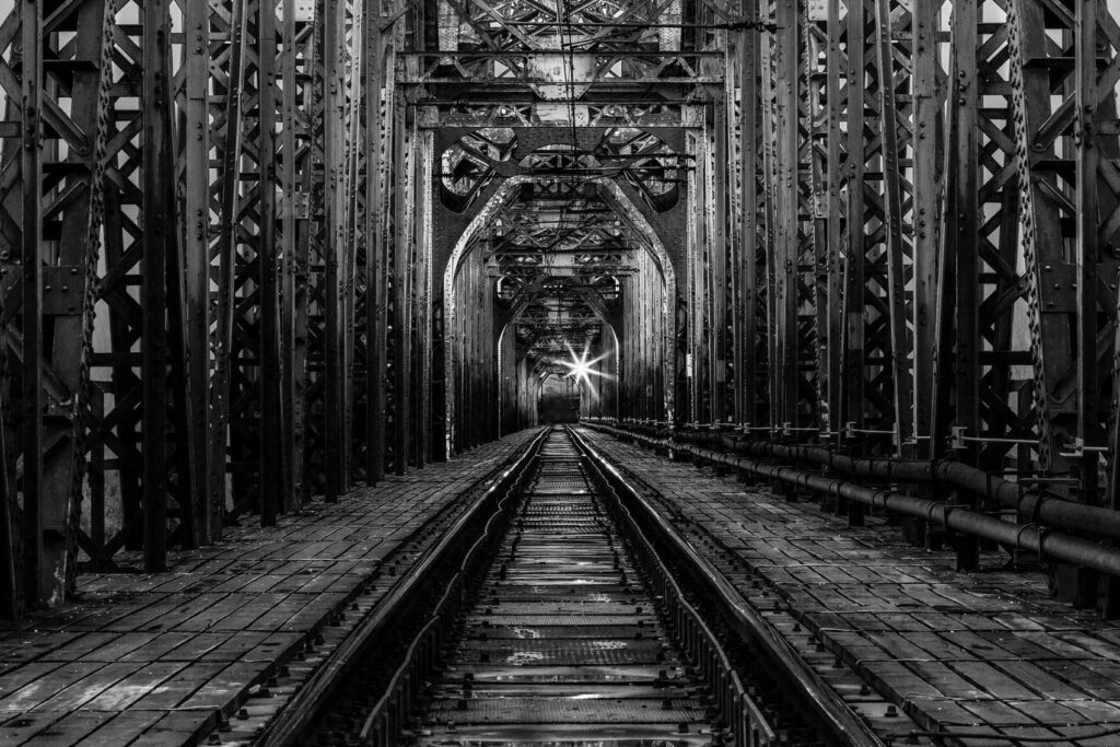 Czarno-białe zdjęcie stalowego mostu kolejowego z widocznymi torami i światłem na końcu konstrukcji | fot. Marcin Skrobisz