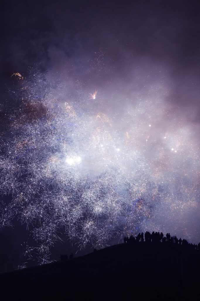 Sylwetki ludzi na wzgórzu obserwujących rozświetlone fajerwerkami nocne niebo | fot. Marcin Skrobisz