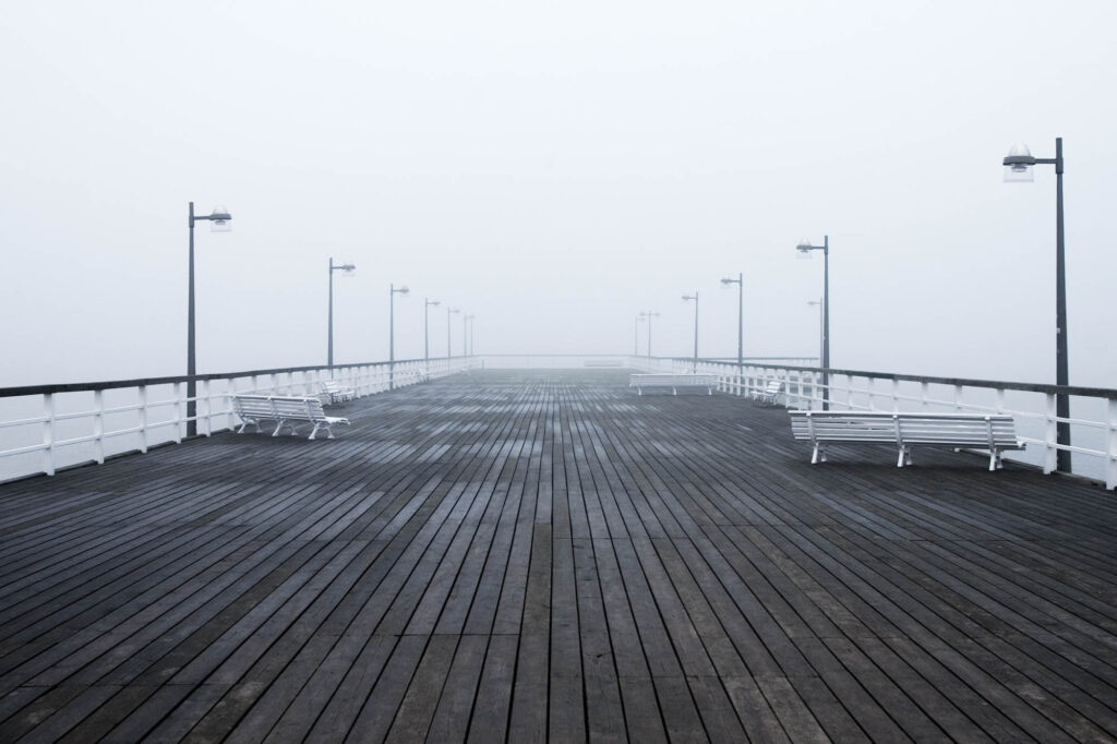 Drewniany pomost z białymi ławkami we mgle, prowadzący w pustkę nad morzem | fot. Marcin Skrobisz